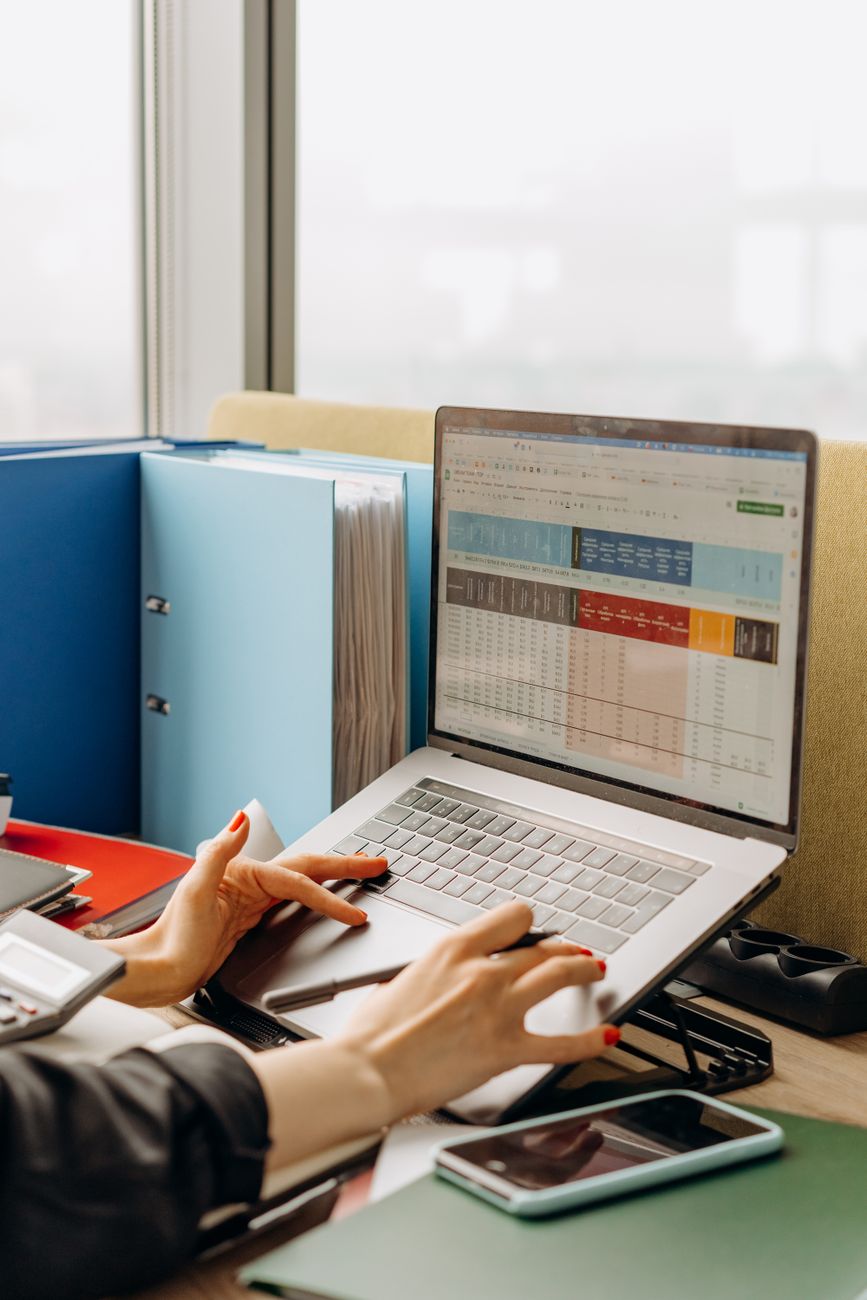 Hands working through a spreadsheet of figures on a laptop beside ring binders